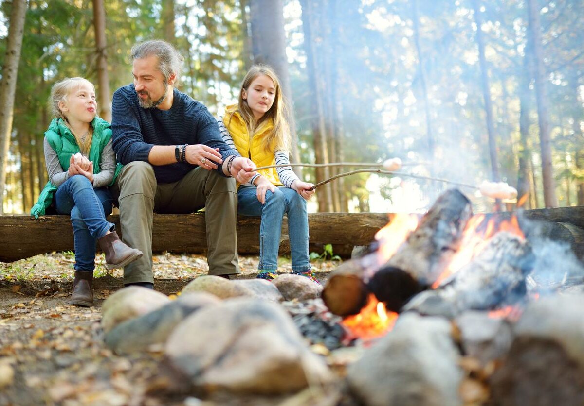 sisters and their father roasting marshmallows on sticks at bonfire.