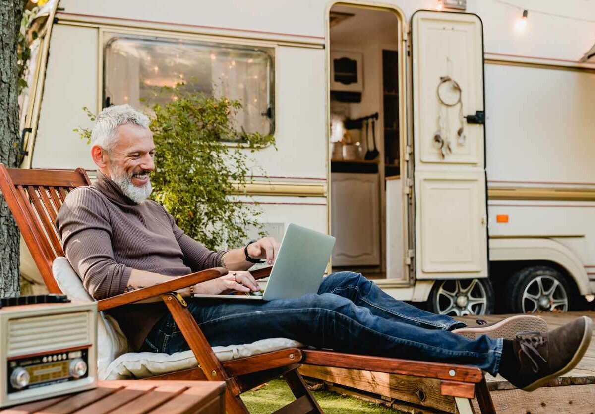 A man with white hair and a white beard relaxes on a wooden chair outside of a white RV with the door open.