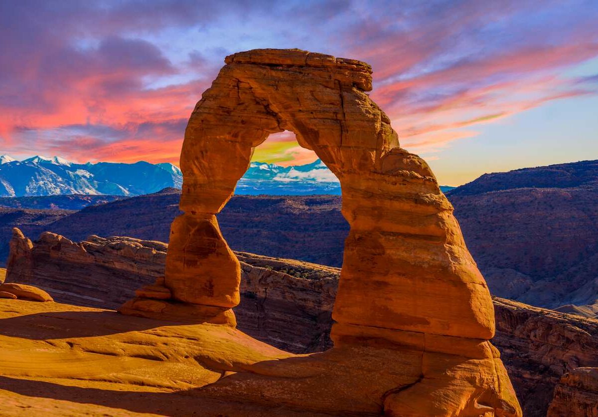 The sun sets over an orange stone arch at Arches National Park, with a pink, orange, and blue sky in the background.