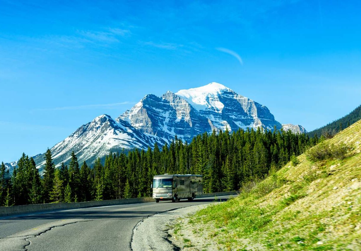An RV goes up a road with the mountains and neighboring forest in the background.